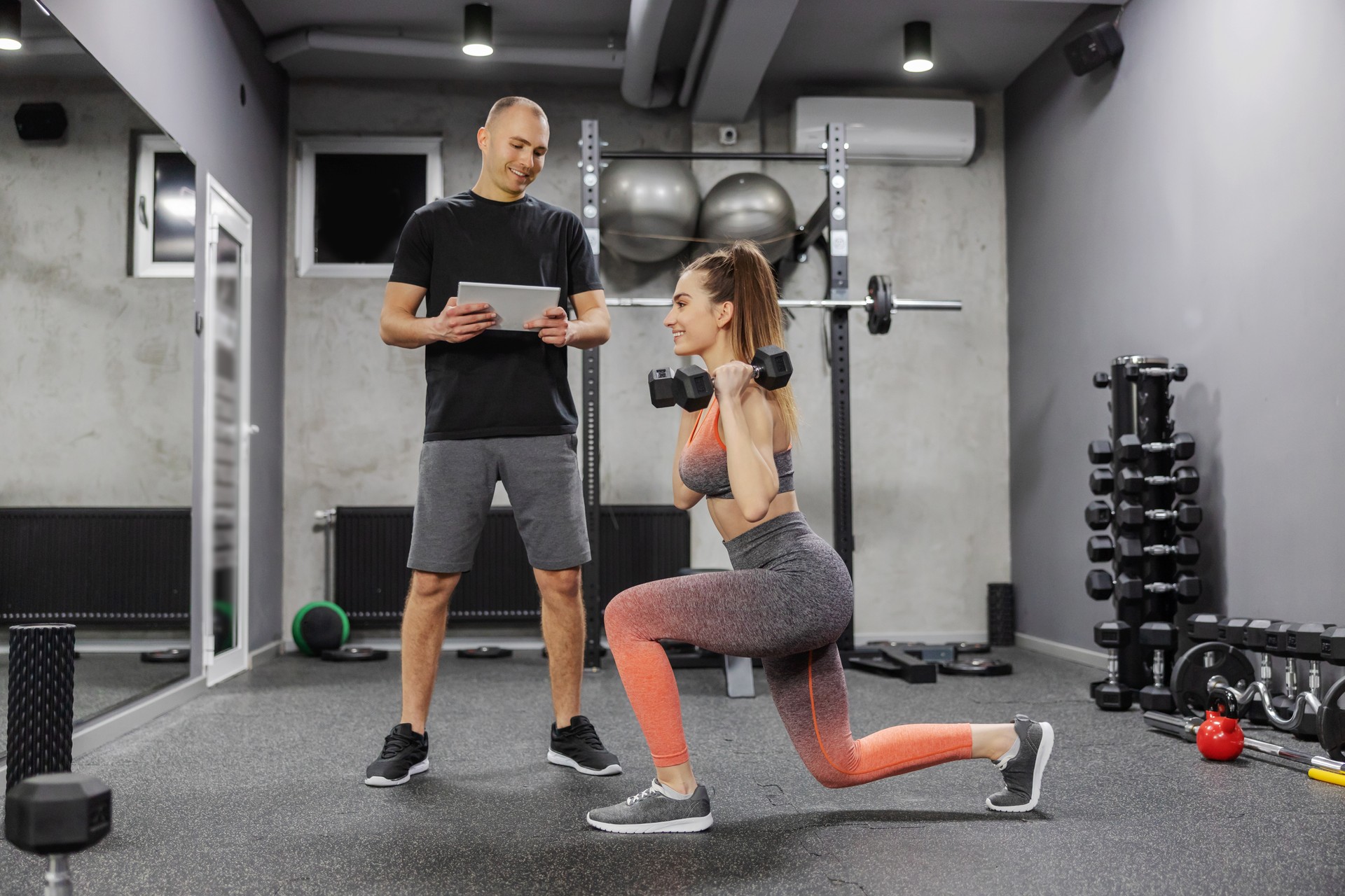 Measuring the success of fitness training Woman in sportswear makes a step forward in the gym and holds dumbbells in her hands while a coach observes her progress and enters data into a digital tablet